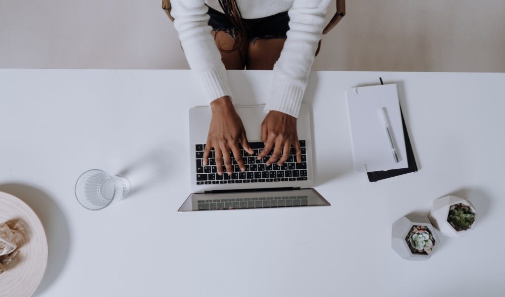 A woman wearing a white sweater is writing her first blog post on a laptop and has a notebook and a glass of water next to her