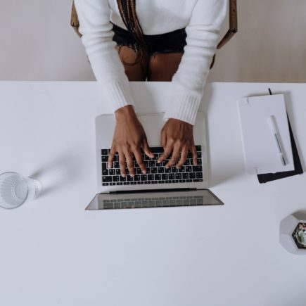 A woman wearing a white sweater is writing her first blog post on a laptop and has a notebook and a glass of water next to her