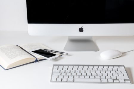 A white desk with a keyboard, a mouse, a monitor, an open book with a phone on top with resources about how to start a blog